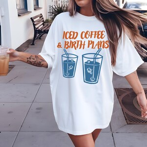 Puede incluir: Camiseta blanca con el texto naranja "ICED COFFEE & BIRTH PLANS" sobre dos ilustraciones azules de caf&eacute; helado. La camiseta es de estilo casual y holgado.