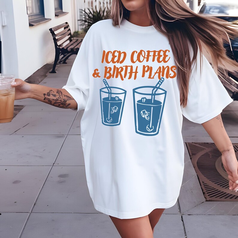 Puede incluir: Camiseta blanca con el texto naranja "ICED COFFEE & BIRTH PLANS" sobre dos ilustraciones azules de caf&eacute; helado. La camiseta es de estilo casual y holgado.
