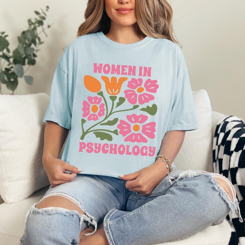 K&ouml;nnte beinhalten: Ein hellblaues T-Shirt mit einem Blumendesign und dem Text "Women in Psychology" auf der Vorderseite.