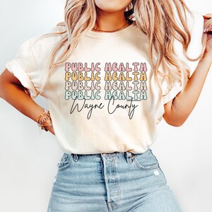 May include: Cream-colored t-shirt with the words "PUBLIC HEALTH" stacked four times in pastel colors, and "Wayne County" in a cursive font. The shirt is paired with blue denim jeans.