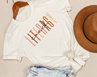 May include: White t-shirt with the text "Literacy Coach" printed in vertical, colorful letters. The letters are in shades of pink, orange, brown, and peach.