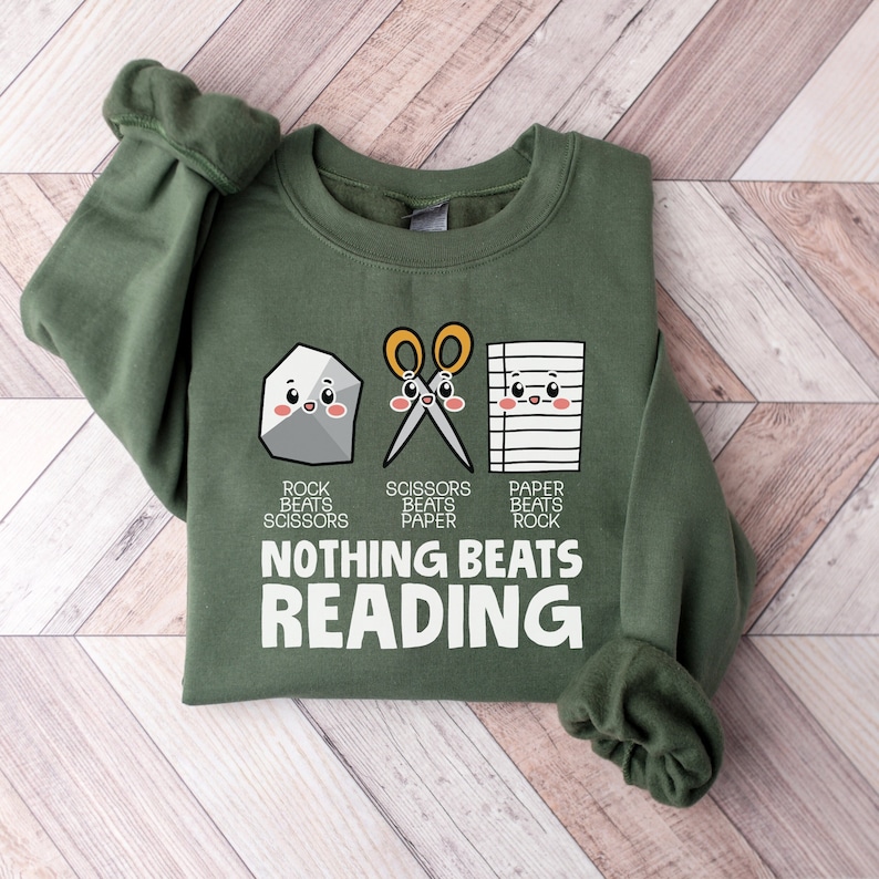 Puede incluir: Una sudadera verde con un gr&aacute;fico de una piedra, unas tijeras y un papel con el texto "Piedra vence a tijeras, tijeras vence a papel, papel vence a piedra, nada vence a la lectura".