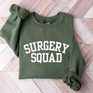 May include: A green sweatshirt with the text "SURGERY SQUAD" in white letters.