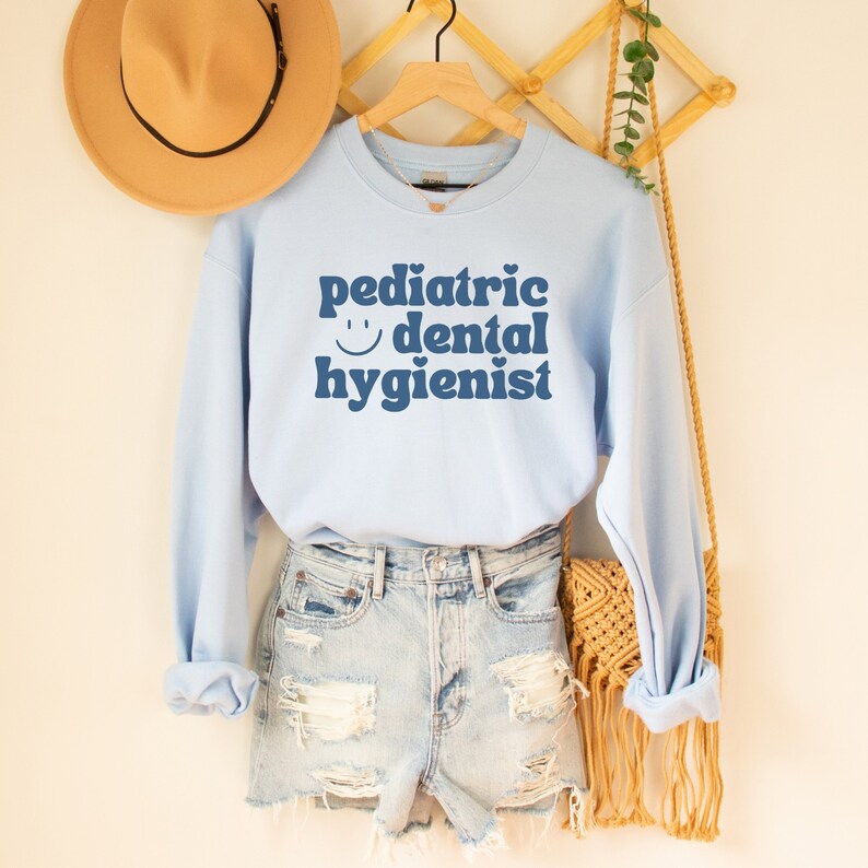 May include: Light blue sweatshirt with the text "pediatric dental hygienist" in blue with a smiley face and a heart. The sweatshirt is on a hanger with a straw hat and a woven bag.