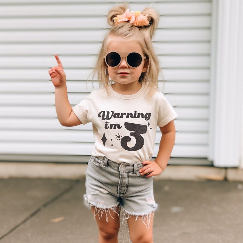 May include: A young child wearing a light beige t-shirt with the text "Warning I'm 3" in black. The child is wearing round sunglasses and has their hair pulled back in two pigtails with a pink flower.