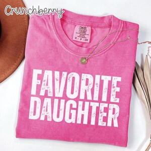 May include: A pink short-sleeved t-shirt with a crew neckline and the text "FAVORITE DAUGHTER" printed in large white distressed capital letters. The shirt features a Comfort Colors brand label on the inside collar with size S P CH.