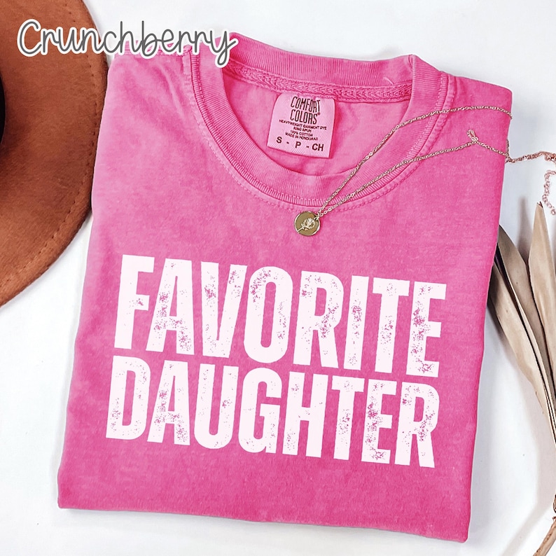 May include: A pink short-sleeved t-shirt with a crew neckline and the text "FAVORITE DAUGHTER" printed in large white distressed capital letters. The shirt features a Comfort Colors brand label on the inside collar with size S P CH.