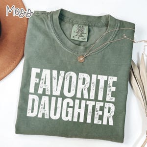 May include: A moss green crew neck t-shirt featuring the text FAVORITE DAUGHTER in distressed white capital letters. The shirt has a Comfort Colors brand label at the inner neckline and is paired with a thin gold chain necklace with a small pendant.