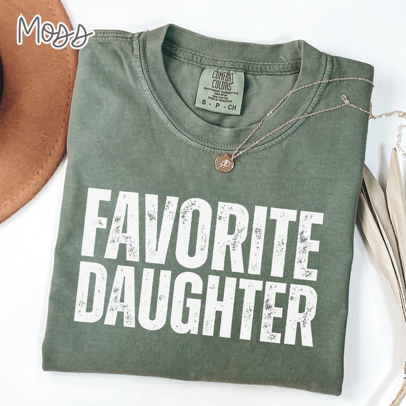 May include: A moss green crew neck t-shirt featuring the text FAVORITE DAUGHTER in distressed white capital letters. The shirt has a Comfort Colors brand label at the inner neckline and is paired with a thin gold chain necklace with a small pendant.