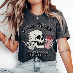May include: Dark gray t-shirt with the text "STILL SOBER BITCHES" above a skull graphic. The skull is holding a pink coffee cup that says "sober". The skull is giving the middle finger with a skeleton hand.