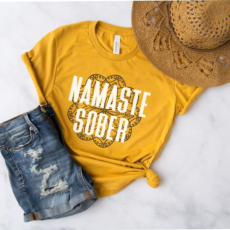 May include: Mustard yellow t-shirt with the text "NAMASTE SOBER" in white, set against a black mandala design. The shirt is paired with denim shorts and a straw hat. The shirt is knotted at the waist.