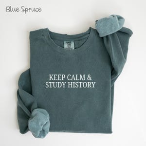 May include: A blue spruce sweatshirt with the text "KEEP CALM & STUDY HISTORY" in white. The long-sleeved shirt is folded, with the sleeves bunched up. The text "Blue Spruce" is in the upper left corner.