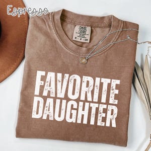 May include: A brown short-sleeved t-shirt with the text "FAVORITE DAUGHTER" printed in white distressed lettering on the front. The shirt features a crew neckline and a Comfort Colors brand label inside the collar. A silver chain necklace is present.