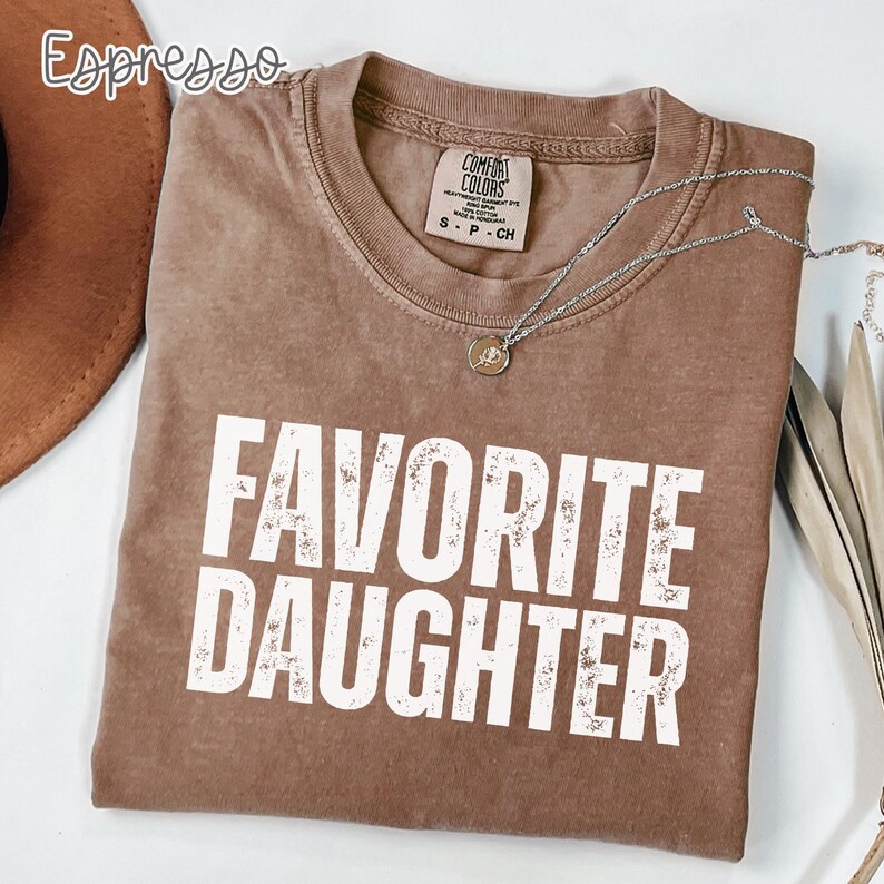 May include: A brown short-sleeved t-shirt with the text "FAVORITE DAUGHTER" printed in white distressed lettering on the front. The shirt features a crew neckline and a Comfort Colors brand label inside the collar. A silver chain necklace is present.
