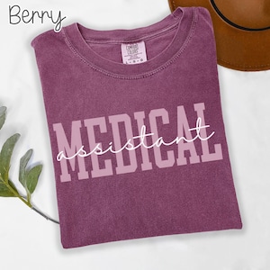 May include: Berry-colored t-shirt with the words "MEDICAL assistant" printed in a light purple color. The shirt is a Comfort Colors brand and has a crew neck. A brown hat is visible in the background.