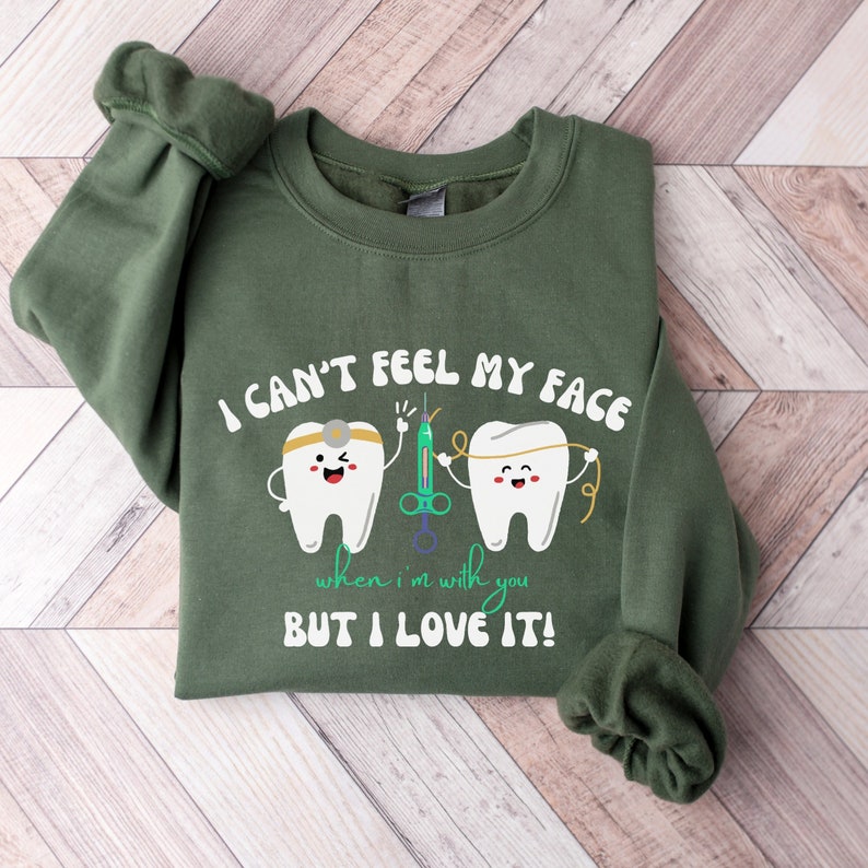 May include: Green sweatshirt with the text "I CAN'T FEEL MY FACE when I'm with you BUT I LOVE IT!". The design features two cartoon teeth, a syringe, and dental floss. The sweatshirt is folded on a wooden surface.