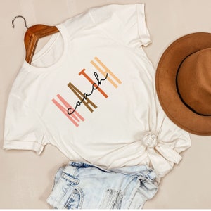 May include: White t-shirt with the text "MATH coach" in black script with colorful stripes behind the letters.