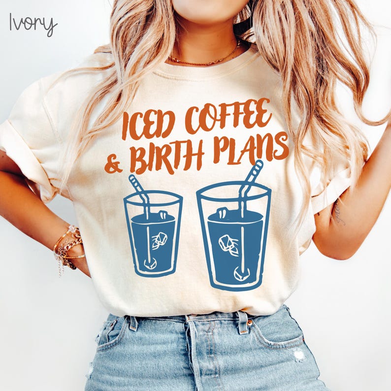 Puede incluir: Camiseta color marfil con el texto naranja "ICED COFFEE & BIRTH PLANS". Debajo, dos vasos azules de caf&eacute; helado con pajitas y cubitos de hielo. La camiseta es de estilo casual.