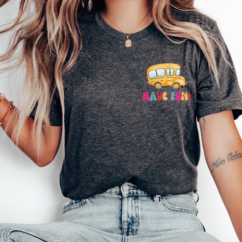 May include: A dark grey t-shirt featuring a yellow school bus graphic and the text "HAVE FUN!" in colourful letters.