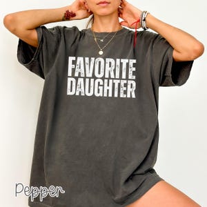 May include: A charcoal grey short-sleeve t-shirt featuring the white distressed text FAVORITE DAUGHTER printed in a bold, sans-serif font across the chest. The garment has a crew neckline and a relaxed, loose-fitting silhouette.