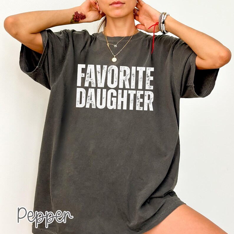 May include: A charcoal grey short-sleeve t-shirt featuring the white distressed text FAVORITE DAUGHTER printed in a bold, sans-serif font across the chest. The garment has a crew neckline and a relaxed, loose-fitting silhouette.