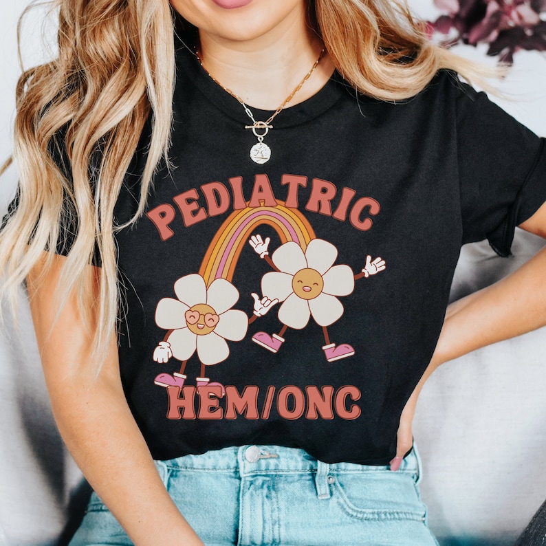 Peut inclure: T-shirt noir avec un arc-en-ciel et deux fleurs de dessin anim&eacute;. Le texte "PEDIATRIC HEM/ONC" est imprim&eacute; sur le t-shirt.