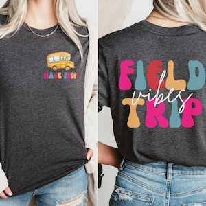 May include: A dark grey t-shirt with a yellow school bus graphic on the front and the text "HAVE FUN!" underneath. The back of the shirt has the text "FIELD vibes TRIP" in colourful, retro-style letters.