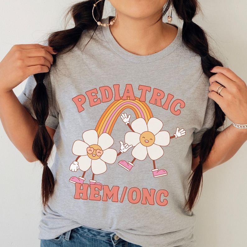 Peut inclure: Un t-shirt gris avec le texte "PEDIATRIC HEM/ONC" en lettres roses et orange. Deux fleurs de dessin anim&eacute; avec des visages souriants se tiennent la main devant un arc-en-ciel.