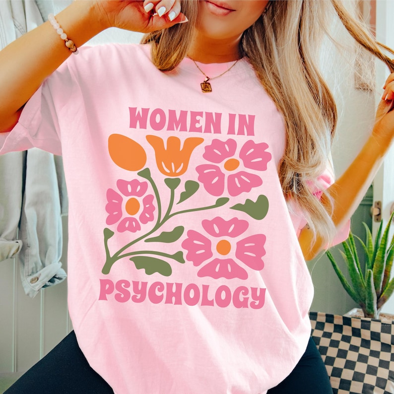K&ouml;nnte beinhalten: Pinkes T-Shirt mit einem floralen Design und dem Text "Women in Psychology" in einer Retro-Schrift.