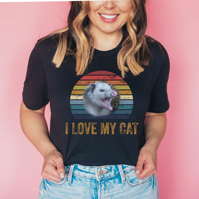 Puede incluir: Una camiseta negra con un gr&aacute;fico de puesta de sol retro que presenta un opossum con el texto "I LOVE MY CAT" en amarillo.