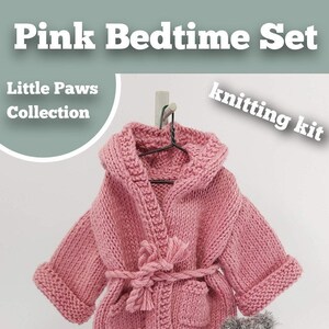 May include: Pink knitted bedtime set knitting kit for a child. The set includes a hooded robe, slippers, a small bag, and a teddy bear. The robe has a tie belt and a pocket. The slippers have white fur trim and pink pom-poms. The bag is pink and has a white lining. The teddy bear is gray and fuzzy.