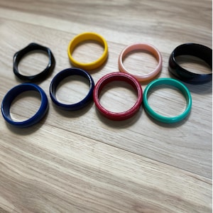May include: A collection of eight circular bangle bracelets in various solid colors including black, yellow, light pink, dark blue, red, and teal. Each bracelet features a smooth, rounded band design with a consistent thickness and diameter.