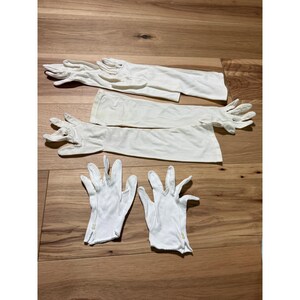 May include: A collection of white gloves consisting of two pairs of long opera-length gloves and one pair of short wrist-length gloves. The short gloves feature small white buttons at the wrist, while the long gloves extend past the elbow.