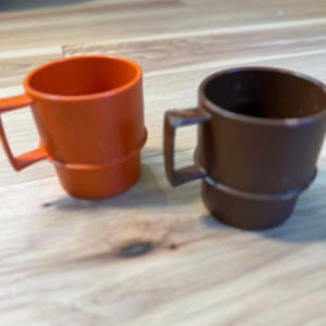 May include: Two stackable mugs, one in a vibrant orange and the other in a deep brown, each featuring a distinct horizontal ridge around the center and a square-shaped handle. These simple, solid-colored drinking vessels have a smooth finish.