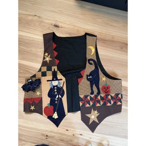 May include: A sleeveless vest featuring a patchwork design with various fabric patterns in black, tan, red, and yellow. Appliqué elements include a witch with a broom, a black cat, a bat, a crescent moon, stars, and a pumpkin. The vest has a solid black back.