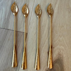 May include: A set of four long-handled gold-toned metal spoons. Each spoon features a bowl shaped like a leaf with etched vein details and a slender stem that widens toward the base, which is also decorated with an etched leaf pattern.