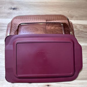 May include: A rectangular amber-tinted glass baking dish with a matching deep red plastic lid. The lid features the embossed Pyrex logo in the center. The dish has integrated handles on the sides and a smooth, transparent glass construction.