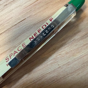 May include: A clear plastic ballpoint pen with a green cap and a silver metal pocket clip. The barrel features the red text SPACE NEEDLE printed vertically, and the metal clip is embossed with the word SEATTLE in a stylized font.