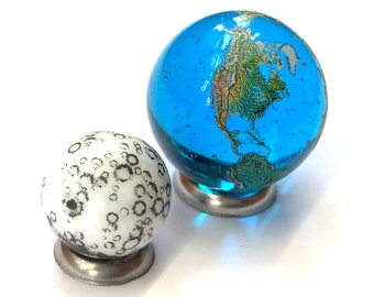 Peut inclure: Deux billes en verre, l'une bleue avec une carte du monde et l'autre blanche avec un motif noir et blanc, sur des supports en métal.