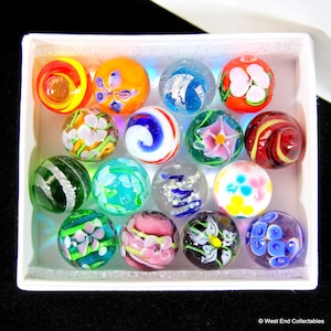 May include: A collection of 15 colorful glass marbles in a white box. The marbles feature various designs, including swirls, flowers, and stripes.