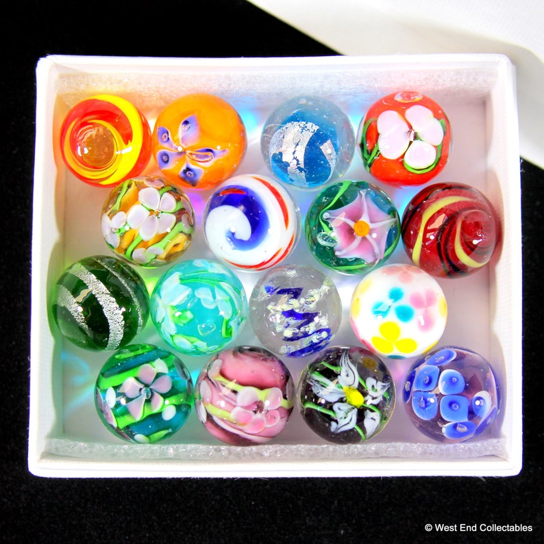May include: A collection of 15 colorful glass marbles in a white box. The marbles feature various designs, including swirls, flowers, and stripes.