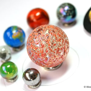 Może przedstawiać: Miniaturowy model Układu Słonecznego z szklanymi kulkami reprezentującymi planety. Planety są ułożone w okrągły wz&oacute;r na białym tle. Planety są pomalowane na r&oacute;żne kolory, w tym niebieski, zielony, czerwony i pomarańczowy. Słońce jest reprezentowane przez ż&oacute;łtą kulkę z czarną podstawą.