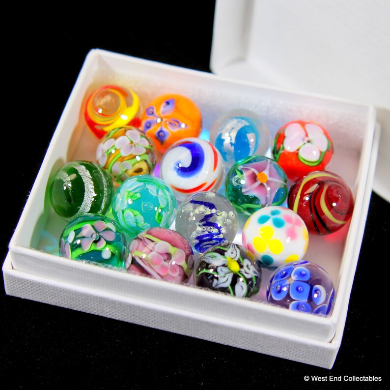 May include: A white box filled with colorful glass marbles. The marbles have various patterns and designs, including flowers, swirls, and stripes.