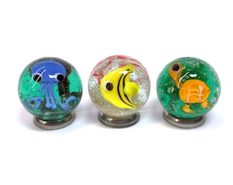 May include: Three glass marbles with different sea creatures inside. The first marble has a blue octopus, the second has a yellow fish, and the third has an orange turtle. All three marbles are on a white background.