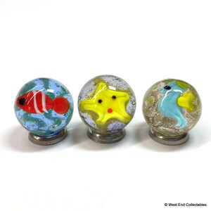 May include: Three glass marbles with a sea life theme. The first marble has a red and white fish with blue and green swirls. The second marble has a yellow starfish with a red center. The third marble has a blue seahorse with yellow and green swirls.
