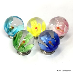 May include: Five glass marbles, each with a different coloured floral design. The colours include blue, yellow, pink, green, and blue. Each marble has a flower with a red centre and sits on a small metal base. The marbles are clear with the floral designs inside.
