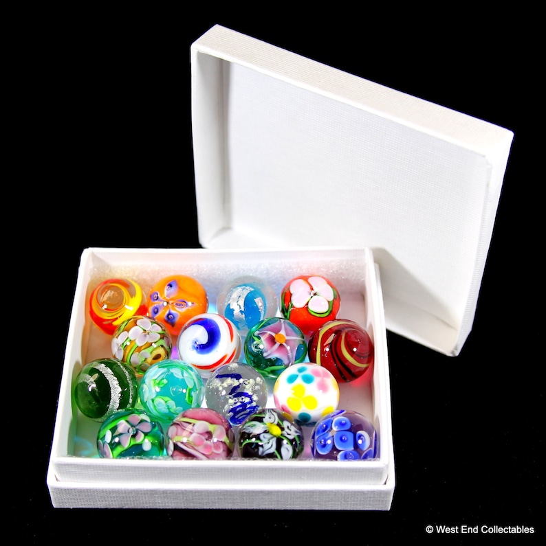 May include: A white box with a lid open, revealing a collection of 16 colorful glass marbles with intricate floral designs. The marbles are arranged in a single layer in the box.