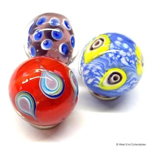 Puede incluir: Tres canicas de vidrio de colores. Una es roja con diseños de gotas azules, blancas y moradas. Otra es azul y amarilla con un patrón similar a un ojo. La tercera es morada con puntos azules.