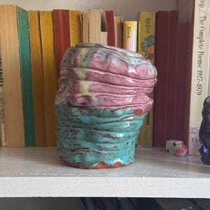 May include: A handmade ceramic vase with a textured, layered design. The vase features bands of pink, turquoise, and yellow glaze. The vase is displayed on a white shelf in front of a row of books.