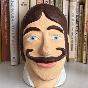 May include: A brown paper mache bust of a man with blue eyes and a brown mustache. The bust is sitting on a shelf with books behind it.